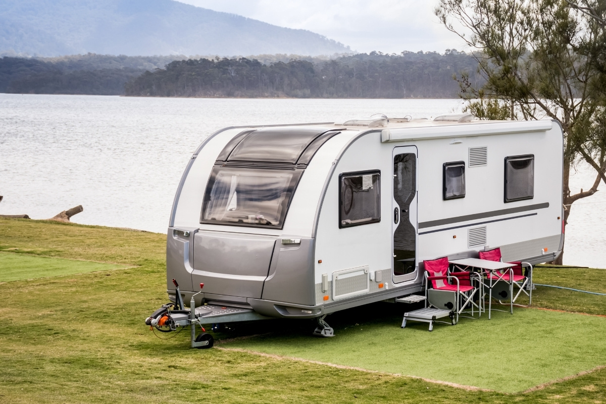 A caravan parked beside a lake, surrounded by trees and reflecting water under a clear blue sky.