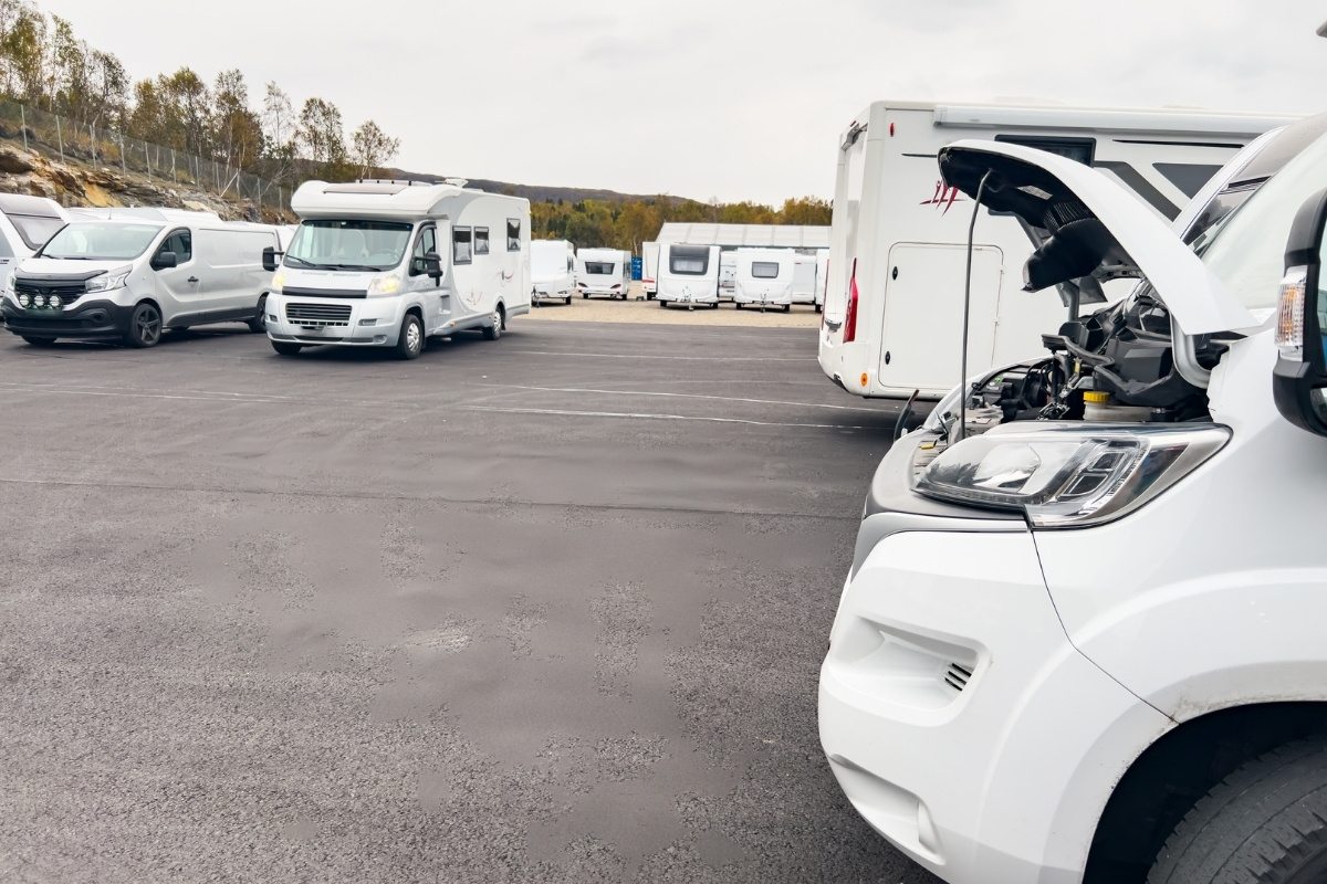 A parking lot with several white camper vans and motorhomes under a cloudy sky. Foreground van has its hood open, suggesting maintenance.
